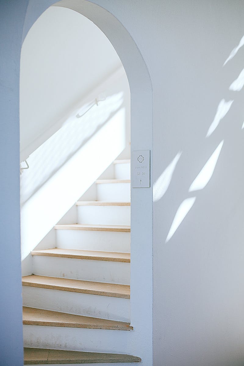 Bright minimalist interior featuring a white staircase with an arch and natural light casting shadows.